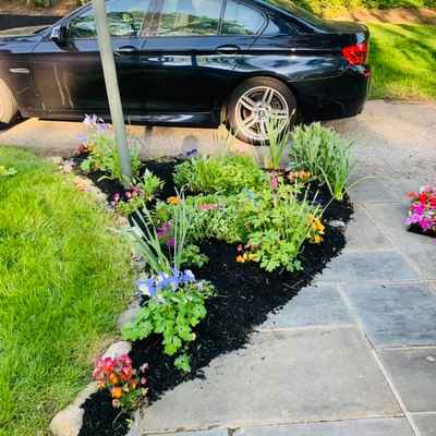A black car is parked on a gravel driveway next to a beautifully designed garden bed, filled with colorful flowers and plants. The artful hardscape design includes a stone pathway leading alongside the garden, bordered by a grassy lawn, all illuminated by daylight.