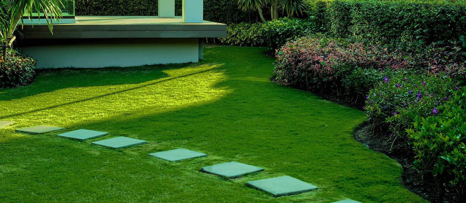 A serene garden with a lush green lawn and a path of square stepping stones leads to a raised wooden deck. Landscapers in Delaware County have ensured the garden is lined with well-maintained hedges and vibrant flowering plants, all bathed in the soft glow of sunlight.