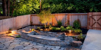 A serene backyard garden features a stone patio and two-tiered stone wall. Soft lighting illuminates surrounding plants and trees, with expert landscapers from Delaware County ensuring the wooden fence in the background complements the lush greenery beyond.