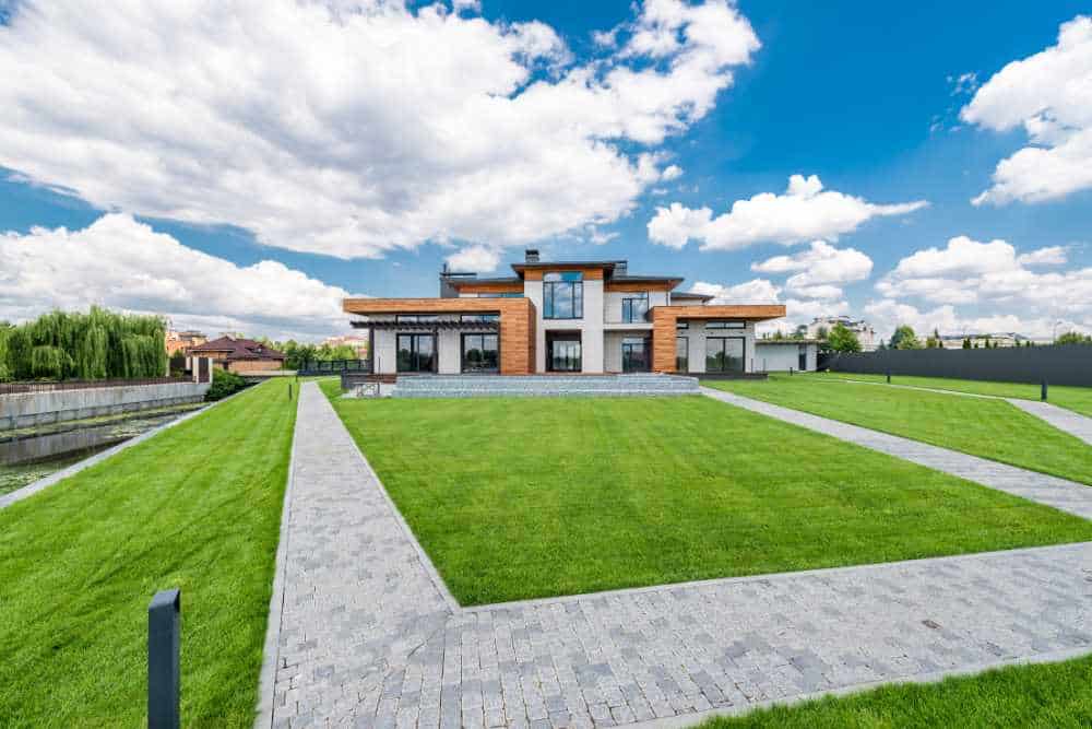 Modern house with large glass windows, surrounded by a manicured lawn and paved pathways, skillfully crafted by landscapers in Delaware County. The sky is blue with scattered clouds. The contemporary design features a blend of wood and stone elements, enhancing its natural elegance.