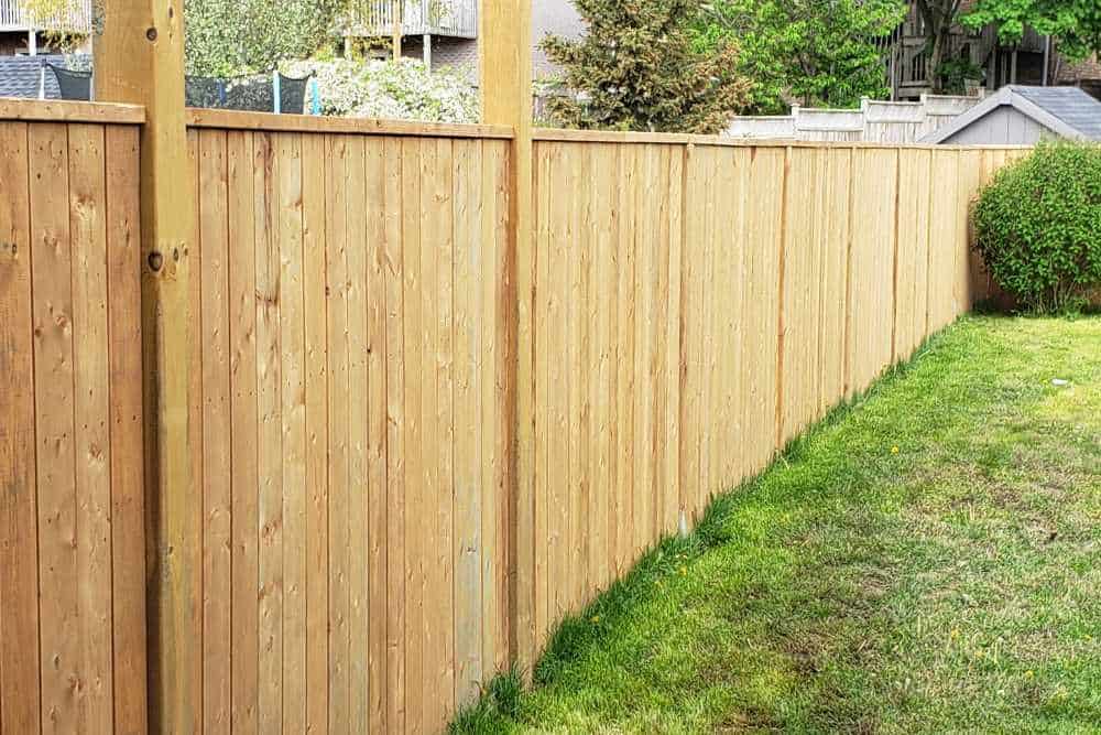 A wooden privacy fence, possibly installed by landscapers in Delaware County, runs the length of a green lawn. In the background, trees and houses are visible. The vertical, evenly spaced panels add a natural touch to the landscape.