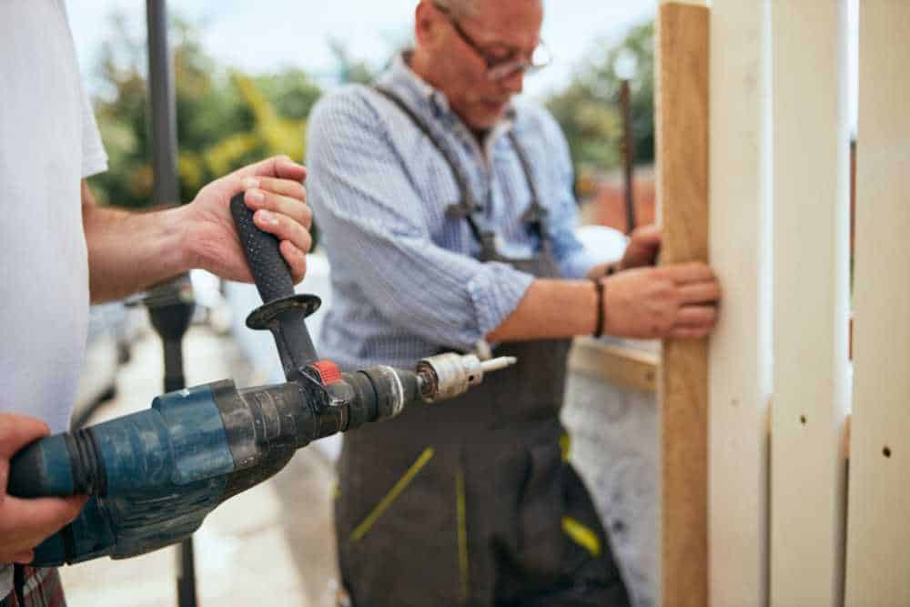 A person using a power drill while another holds wooden planks, possibly for a fence installation in Delaware County. Both are outdoors, wearing casual work clothes. The background is slightly blurred, suggesting a construction or DIY project.