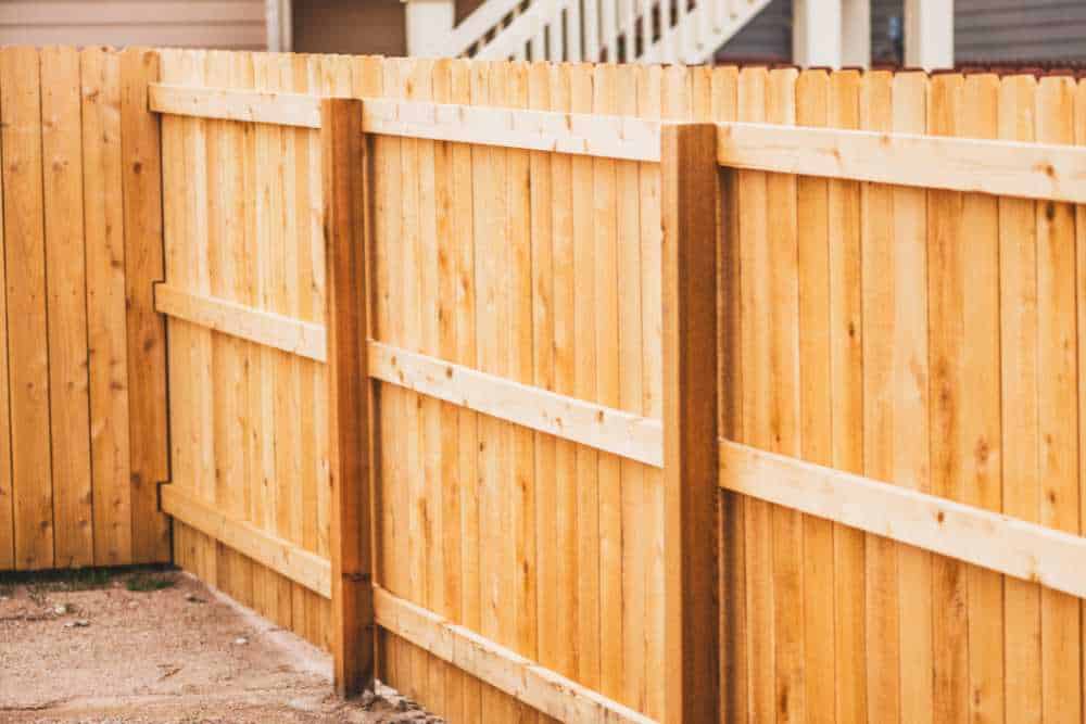 A wooden privacy fence installation in Delaware County, made of light-colored planks, runs along a concrete path. The fence is supported by evenly spaced vertical posts, with a residential building partially visible in the background.