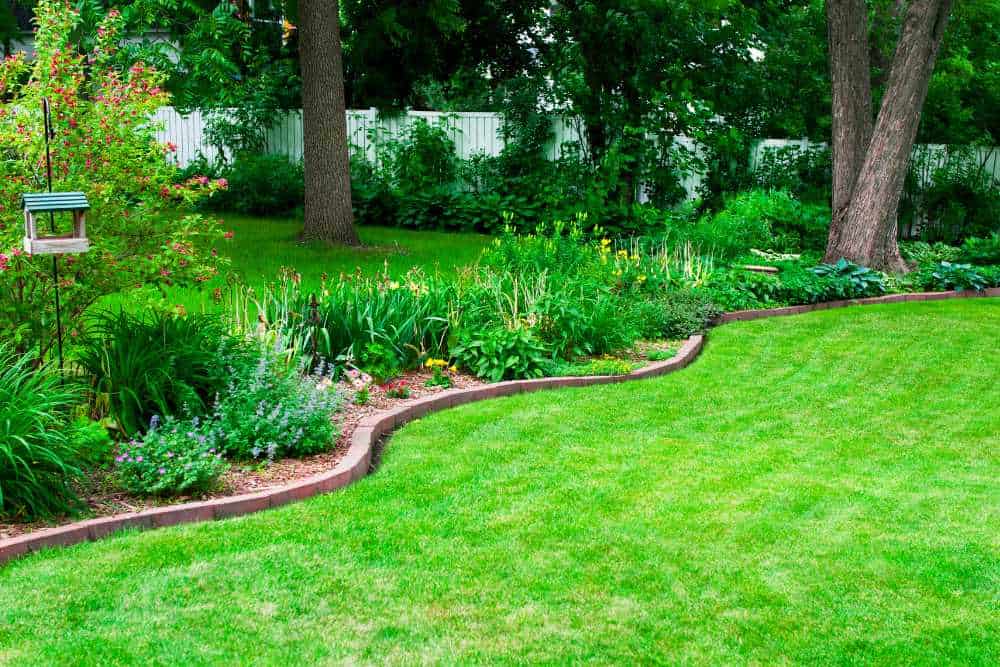 A lush backyard garden with a manicured lawn, bordered by a variety of flowering plants and shrubs, showcases the expert work of landscapers in Delaware County. Tall trees and a white picket fence provide shade in the background, while a small bird feeder hangs on the left.