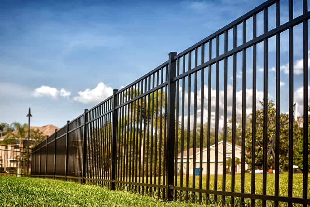 A black metal fence, expertly installed in Delaware County, stretches across a lush green lawn under a bright blue sky with scattered clouds. In the background, houses and trees bask in the sunlight.