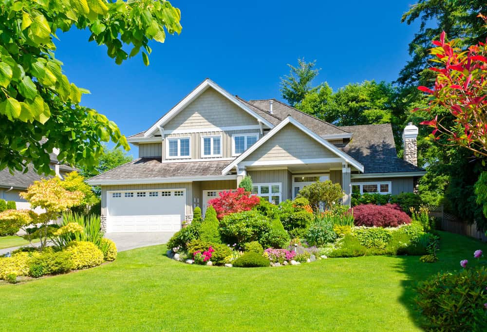 A two-story suburban house with a well-maintained garden featuring colorful flowers and shrubs alongside a lush green lawn. With a gray and white facade, double garage, and new fence installation, it’s surrounded by tall trees under a clear blue sky in Delaware County.