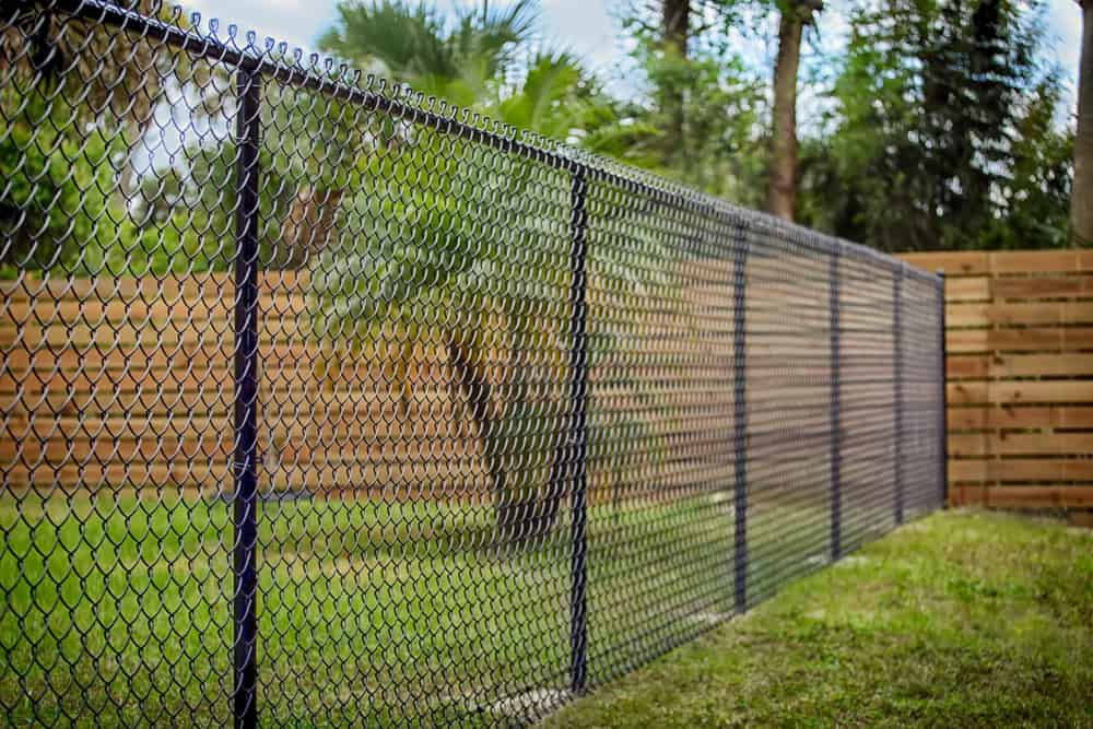 A chain link fence graces the backyard, surrounded by green grass and trees, including a palm tree. A wooden fence runs parallel on the right. The sky is partly cloudy, showcasing the skill of landscapers in Delaware County in creating harmonious outdoor spaces.