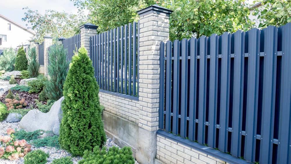 A garden with a landscaped pathway featuring various shrubs and small plants is complemented by a tall, blue metal fence with brick pillars. This tidy area showcases the expertise of landscapers in Delaware County who ensure every detail enhances the overall aesthetic.