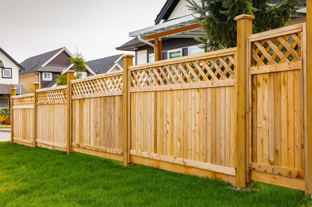 A wooden fence with lattice accents on top stands alongside a row of modern houses. The fence is set on lush green grass, and a large tree is visible behind the fence in a suburban neighborhood.