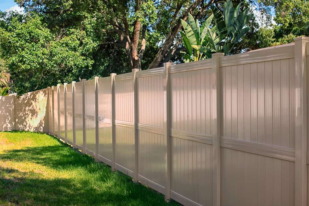 A long, beige vinyl privacy fence borders a grassy area, expertly crafted by landscapers in Delaware County, with tall trees and lush greenery visible in the background under a clear blue sky.
