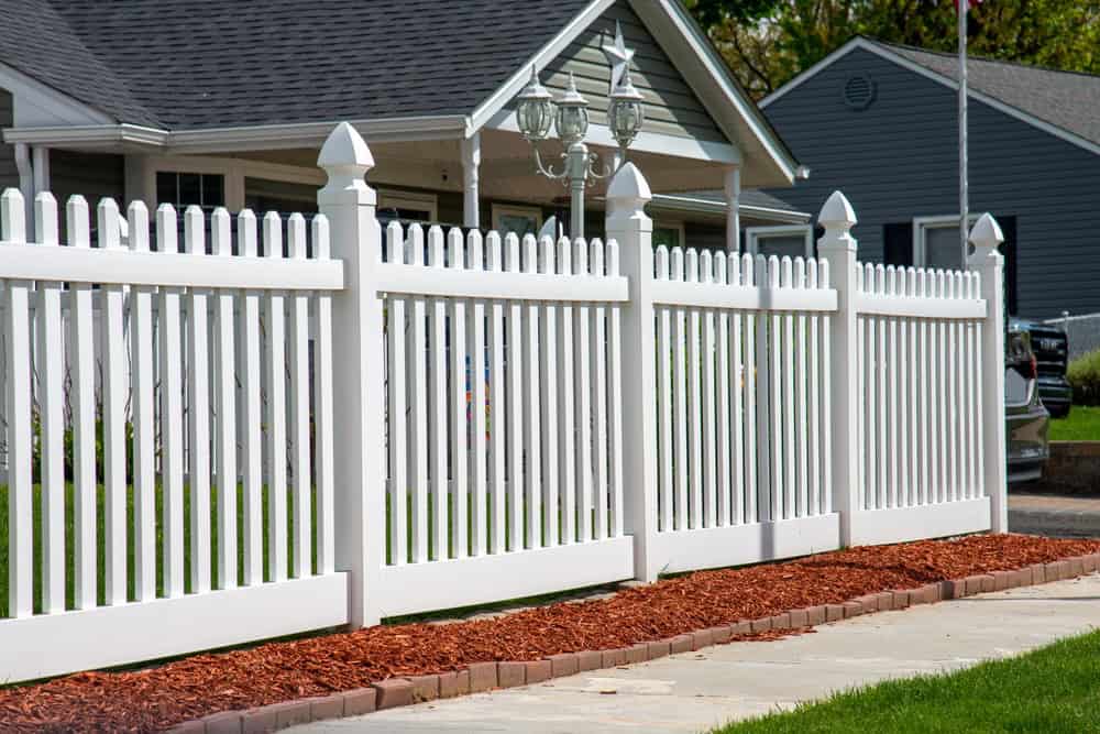 A white picket fence surrounds the front yard of a suburban house. The fence is set on a neatly edged red mulch bed, with green grass visible inside the boundary. A gray house is in the background on a sunny day.