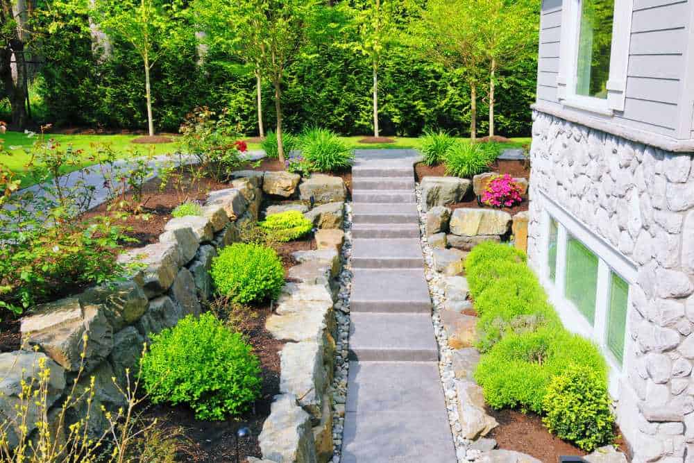 A stone pathway leads to a house, bordered by neatly trimmed bushes and vibrant flowers. The house features a gray stone exterior and several windows. Tall trees and lush greenery encompass the area, ideal for landscapers in Delaware County to add their expert touch.