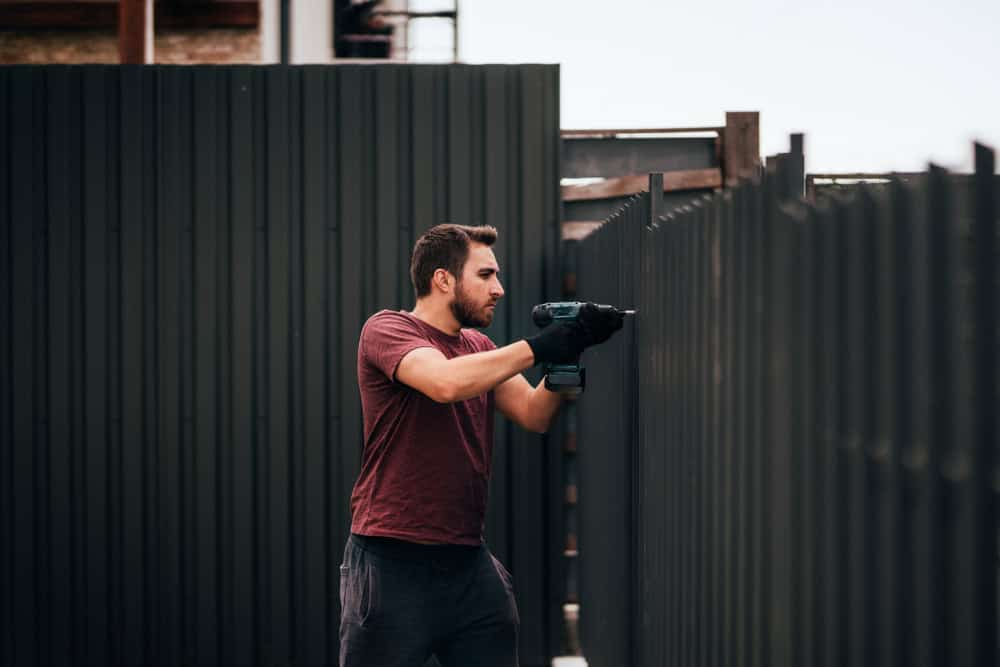 A person in a red shirt and gloves is using a power drill to assemble or repair a tall, dark-colored metal fence outside during the day.