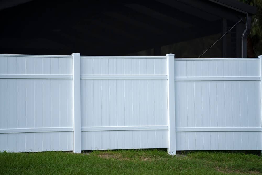 A white vinyl privacy fence with three vertical panels stands on a grassy lawn. Behind the fence, part of a dark structure is visible. The scene is brightly lit, highlighting the clean lines and simple design of the fence.