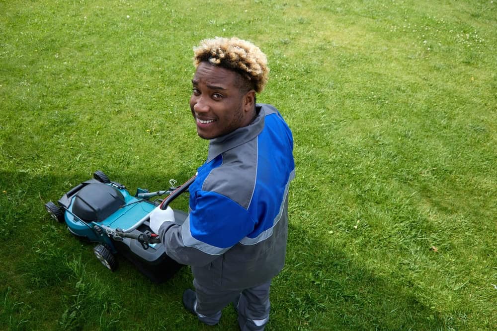 a man in a grey and blue jacket with white gloves and grey pants pushing a turquoise and black lawn mower on a lawn.