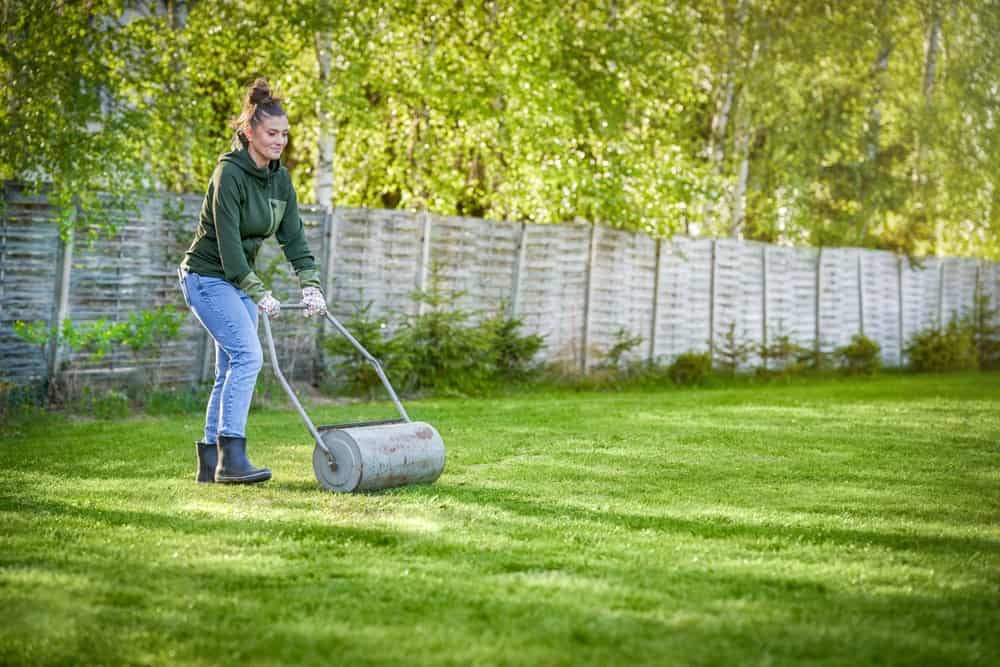 a woman in blue jeans, black boots, and a dark green sweatshirt pushing a grey old fashioned lawn mower on a lush, green lawn.