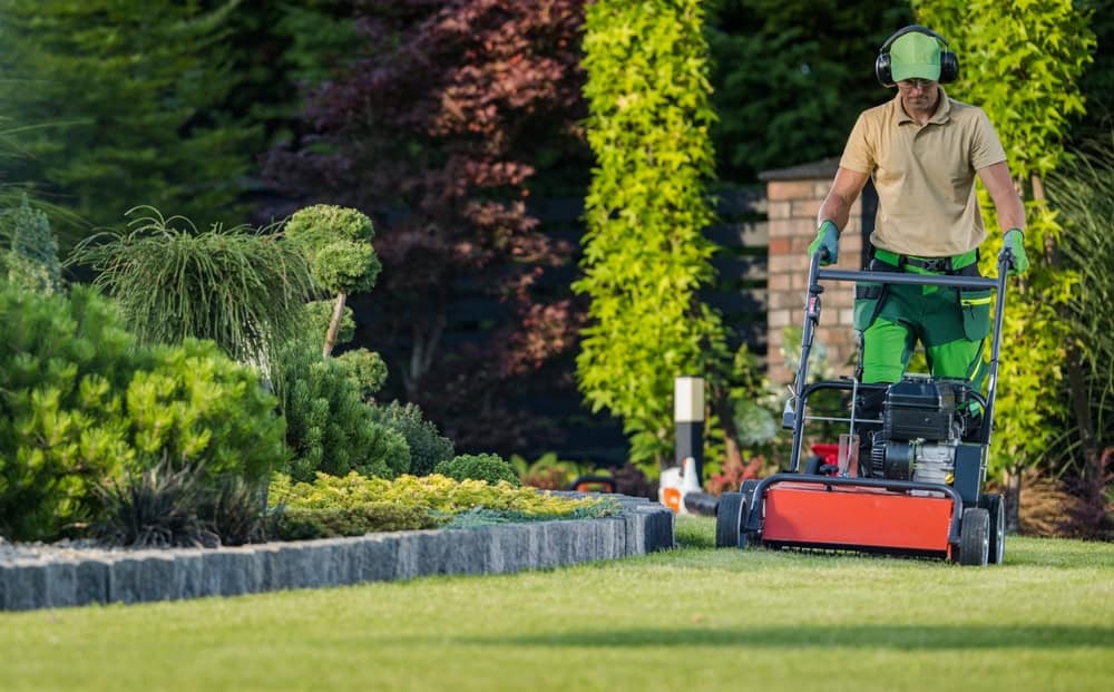 a landscaper in green pants, a tan shirt, green gloves, a green cap, and black headphones using a red lawn mower on a lush lawn. There is a variety of bushes next to him.