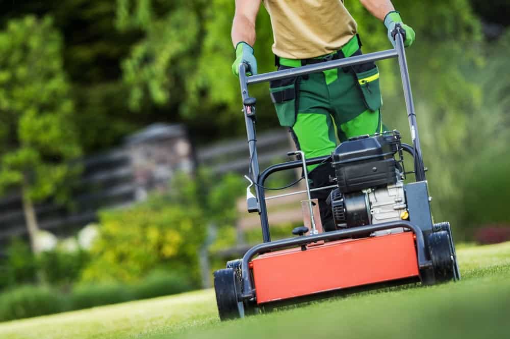 a landscaper in green pants with a tan shirt and green garden gloves using a red lawn mower on a lawn.