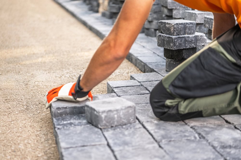A person wearing an orange shirt and work gloves is kneeling and laying gray paving stones in a pattern on a sandy surface. Several stacks of stones are visible nearby, indicating ongoing construction.